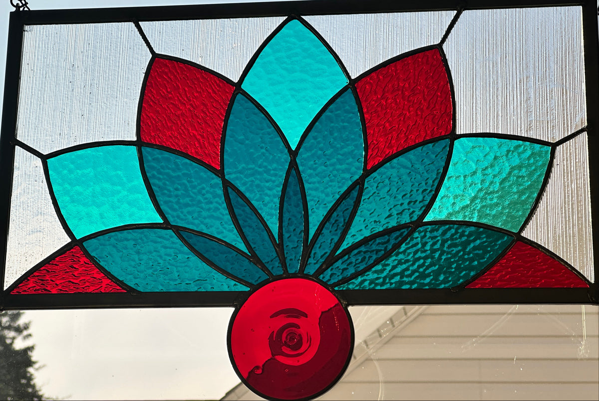 A rectangular stained glass window panel featuring a stylized lotus flower design with red and teal textured glass sections, black metal framing, and hanging chains against an outdoor window background.
